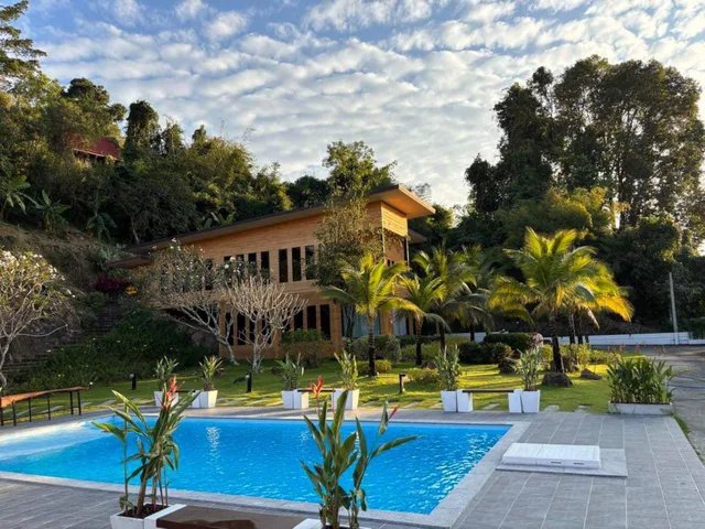 Architect-designed two-storey villa on a green hillside at golden hour, seen across the blue swimming pool with palm trees and tropical garden